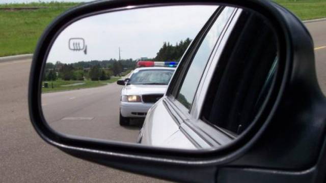 View of police car with flashing lights in rearview mirror on a road, indicating a traffic stop.
