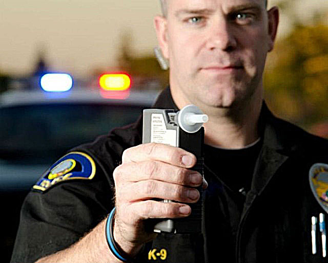Police officer holding a breathalyzer with patrol car in the background, emphasizing safety and law enforcement.