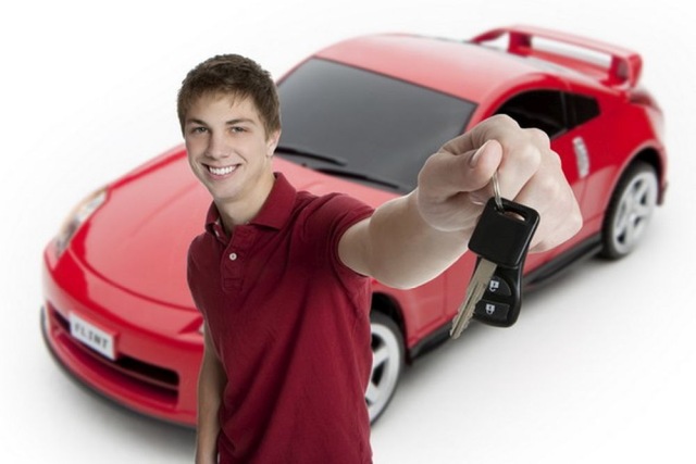 Smiling young man holding car keys in front of a shiny red sports car, symbolizing new car ownership and excitement.