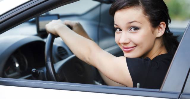 Smiling woman driving a car, looking back over shoulder with a confident expression, hands on the steering wheel.