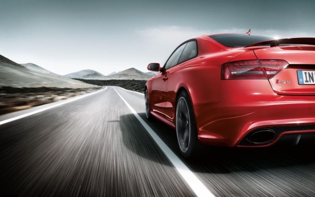Red sports car driving on an open road, mountains in the background, highlighting speed and performance.