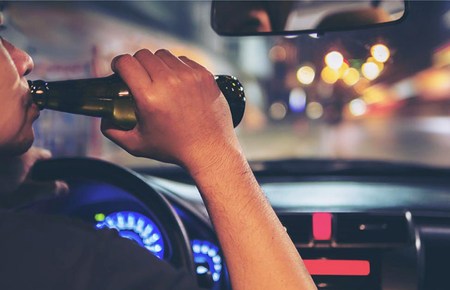 Driver holding a beer bottle while driving at night, highlighting the dangers of drinking and driving.