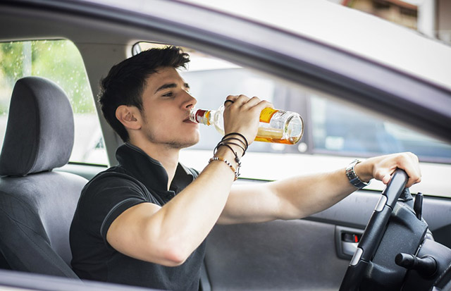 Young man drinking alcohol in driver's seat, highlighting dangers of drinking and driving.
