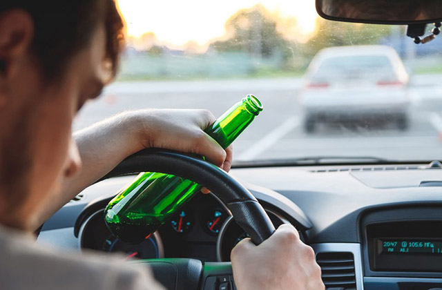 Driver holding beer bottle while driving, emphasizing dangers of drinking and driving.