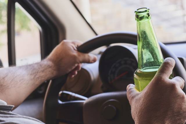Driver holding beer bottle and steering wheel, highlighting the risks of drinking and driving.