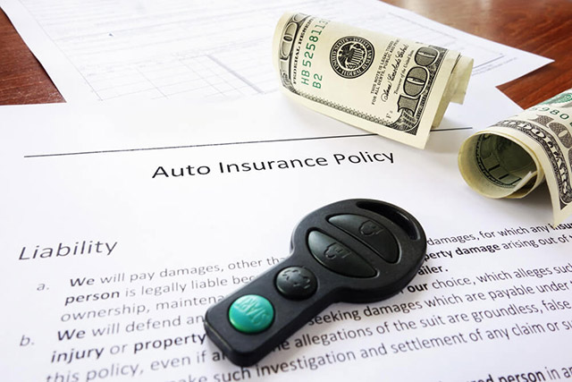 Auto insurance policy document with car key fob and rolled dollar bills on a table, indicating liability coverage.