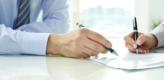 Two people signing a business contract at a desk with pens, wearing formal shirts.