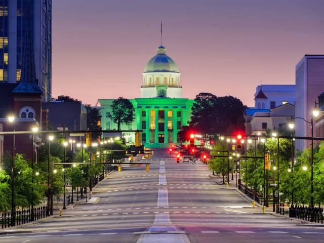 Street view of illuminated Alabama State Capitol building at dusk with vibrant lights and clear sky.