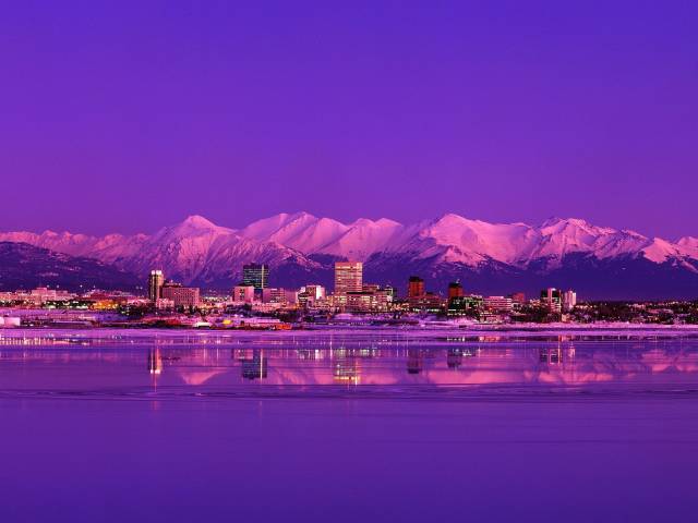 Anchorage skyline at dusk with snow-capped mountains mirrored in serene waters under a vibrant purple sky.