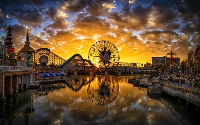 Amusement park sunset with Ferris wheel reflecting in water, vibrant sky, and roller coaster in the background.
