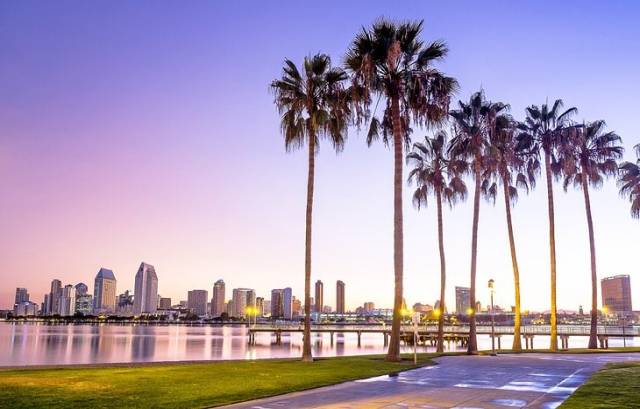 Skyline view of a city at sunset with palm trees in the foreground and a calm body of water reflecting the sky.