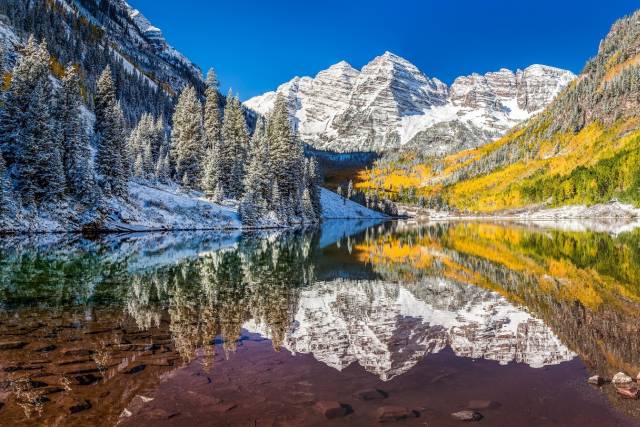 Scenic mountain landscape with snow-capped peaks, colorful autumn trees, and clear lake reflection under a vivid blue sky.
