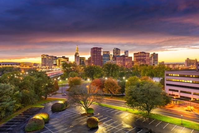 Skyline of Hartford, Connecticut at sunset with colorful sky and illuminated buildings, surrounded by lush green trees.