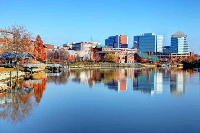 Skyline view of urban buildings reflecting on a calm river under a clear blue sky, showcasing a serene city landscape.