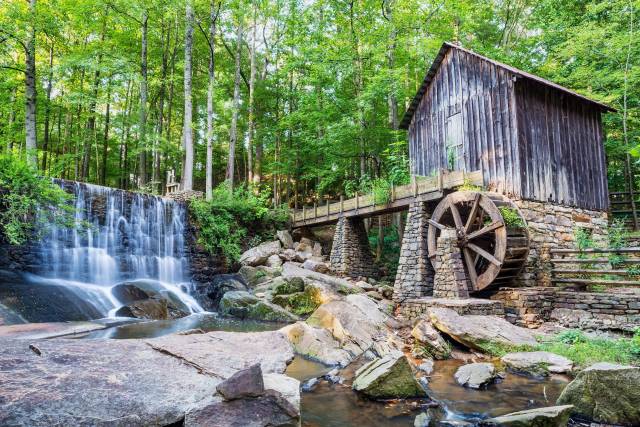 Rustic wooden mill by a waterfall in a lush forest setting, with a stone bridge and waterwheel, under a clear sky.