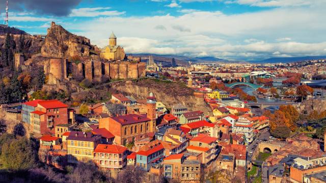 Aerial view of Tbilisi, Georgia with Narikala Fortress and colorful buildings under a cloudy sky.