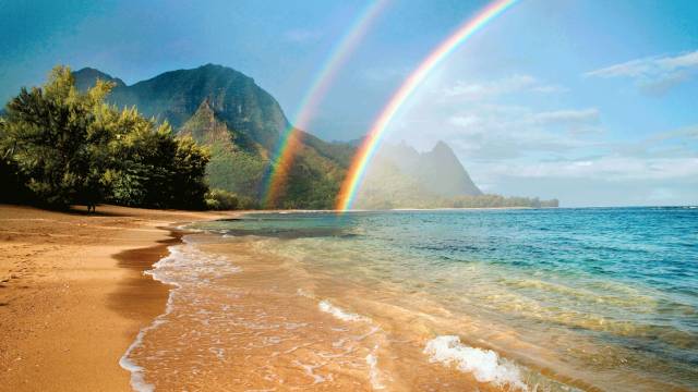 Tropical beach with a vibrant double rainbow over the ocean, lush greenery, and a dramatic mountain backdrop.