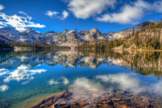 Serene mountain lake reflecting rugged peaks and blue sky, flanked by dense forest, under scattered clouds.