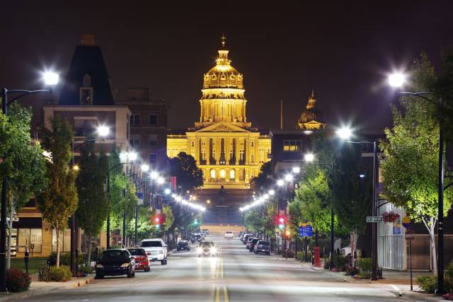 Illuminated capitol building at night with street view and parked cars, trees lining the road in a cityscape setting.
