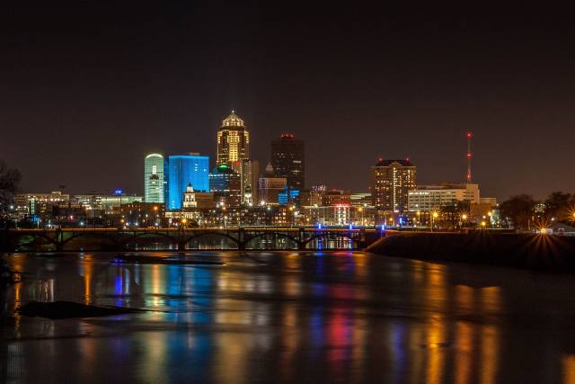 Nighttime skyline of a illuminated city with colorful reflections on the river, featuring prominent buildings and bridge.