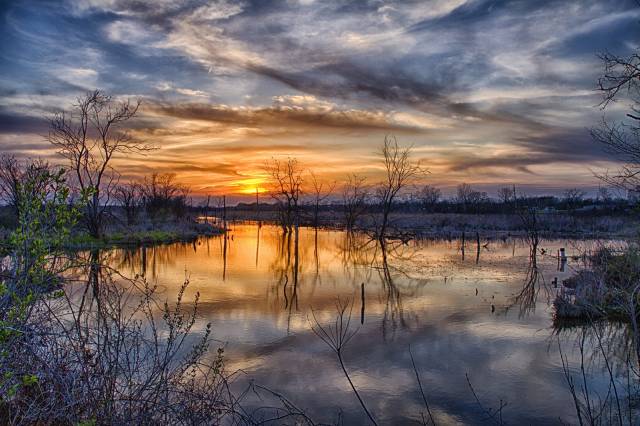 Sunset over a serene lake with reflective water, surrounded by barren trees and a dramatic, colorful sky.