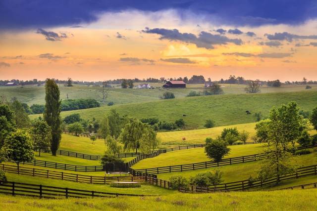 Rolling green hills and fences under a colorful sunset sky, featuring a distant barn and grazing animals.