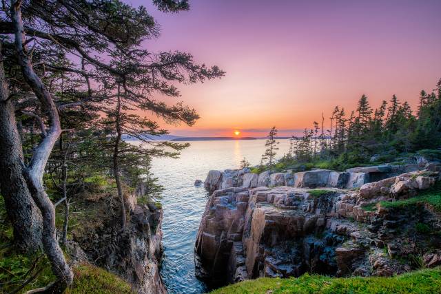 Scenic coastal sunset view with rocky cliffs and pine trees, overlooking a tranquil ocean under a vibrant sky.