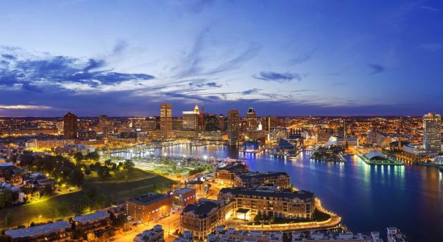 Skyline of Baltimore at dusk, reflecting city lights over the water, with a vibrant sky in the background.