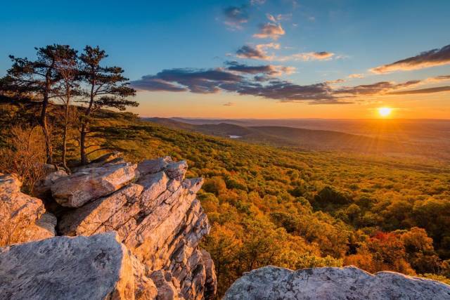 Sunset over Shenandoah Valley from a rocky overlook, featuring trees and vibrant autumn foliage under a colorful sky.
