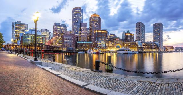 Boston skyline at dusk with waterfront, illuminated skyscrapers, and cloudy sky reflecting in the harbor water.
