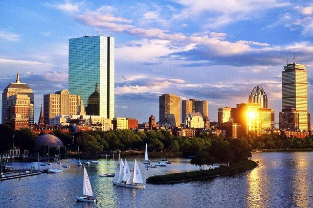 Boston skyline at sunset with sailboats on the Charles River, highlighting city skyscrapers and a vibrant sky.