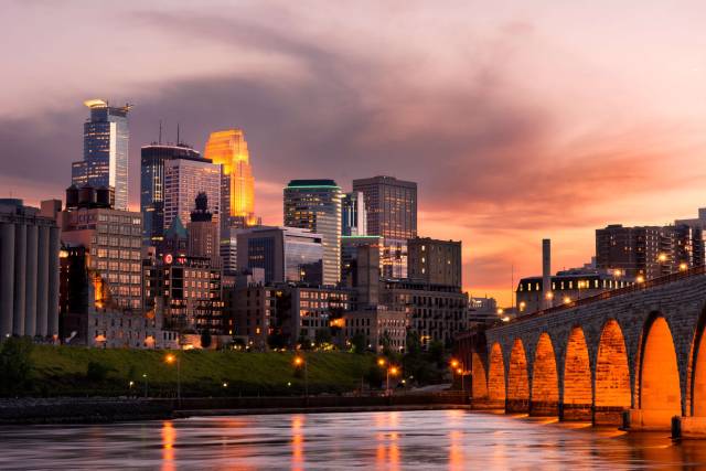 Minneapolis skyline at sunset with Stone Arch Bridge and glowing city lights reflecting on the Mississippi River.