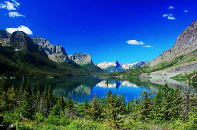 Scenic mountain landscape with a clear lake, lush green forests, and a vibrant blue sky with a few clouds.