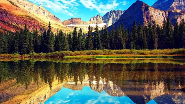 Scenic mountain landscape with evergreen forest reflected in a calm lake under a vibrant blue sky with white clouds.