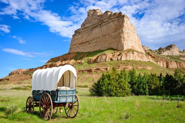 Covered wagon in green field with rocky bluff under blue sky, representing pioneers and outdoor adventures.