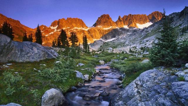 Mountain landscape at sunset with a stream flowing through lush greenery and rocky terrain.