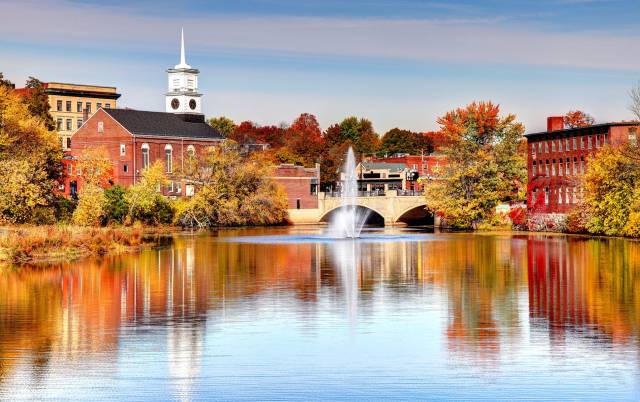 Scenic view of a river reflecting colorful fall foliage and historic buildings with a church steeple and fountain in New England.