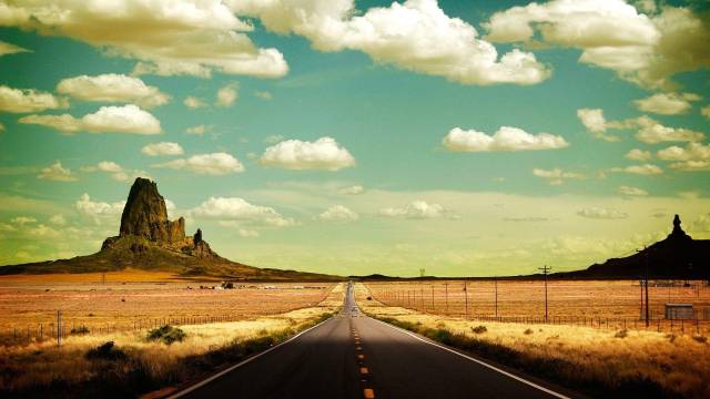 Open desert highway leading to rocky formations under a blue sky with clouds.