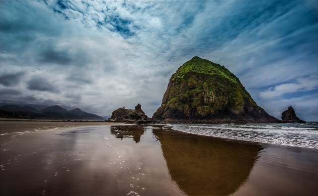 Dramatic coastal scenery with a large rocky outcrop on a beach under a cloudy sky, reflecting in the wet sand.
