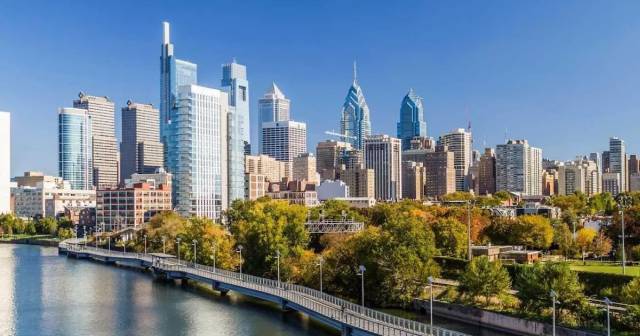 Philadelphia skyline with modern skyscrapers and a river in the foreground under a clear blue sky.