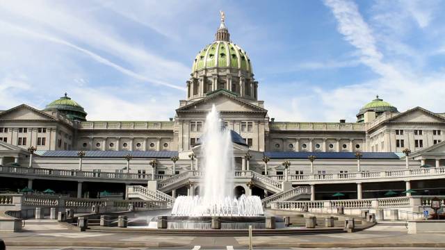 Capitol building with green dome and fountain under a blue sky, showcasing architectural grandeur and historic significance.