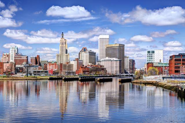 Scenic view of Providence skyline with river reflections under a vibrant blue sky and fluffy white clouds.