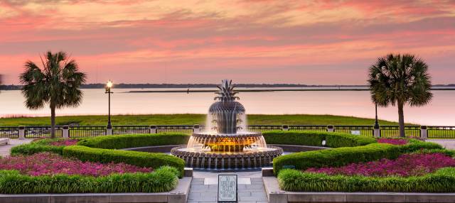 Pineapple Fountain at sunset in Charleston's Waterfront Park, framed by lush greenery and a tranquil waterfront view.