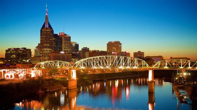 Nashville skyline at sunset featuring the lit bridge over a calm river, showcasing vibrant city lights and architecture.