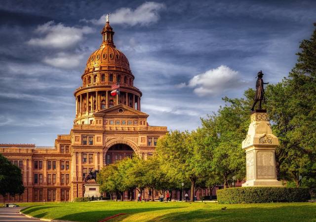 Texas Capitol building under a cloudy sky with a statue and lush greenery in the foreground.