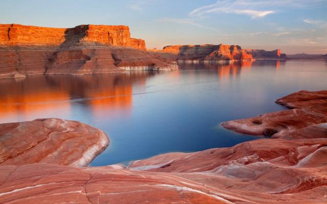 Scenic view of Lake Powell with red rock formations and calm water under a colorful sky at sunset.