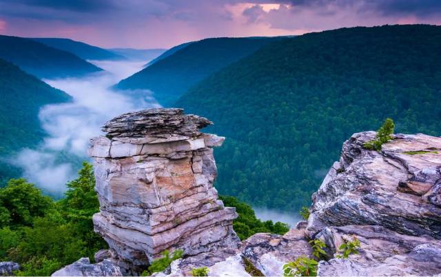 Scenic overlook of a misty valley with lush green hills and rugged rock formations under a purple sky.