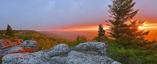 Serene mountain landscape with rocky foreground and vibrant sunset sky, framing lush green forest and horizon.