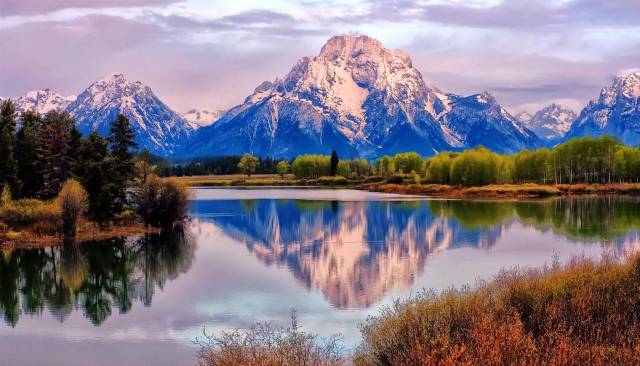 Snowy mountain reflected in a calm lake, surrounded by lush forests under a colorful sky.